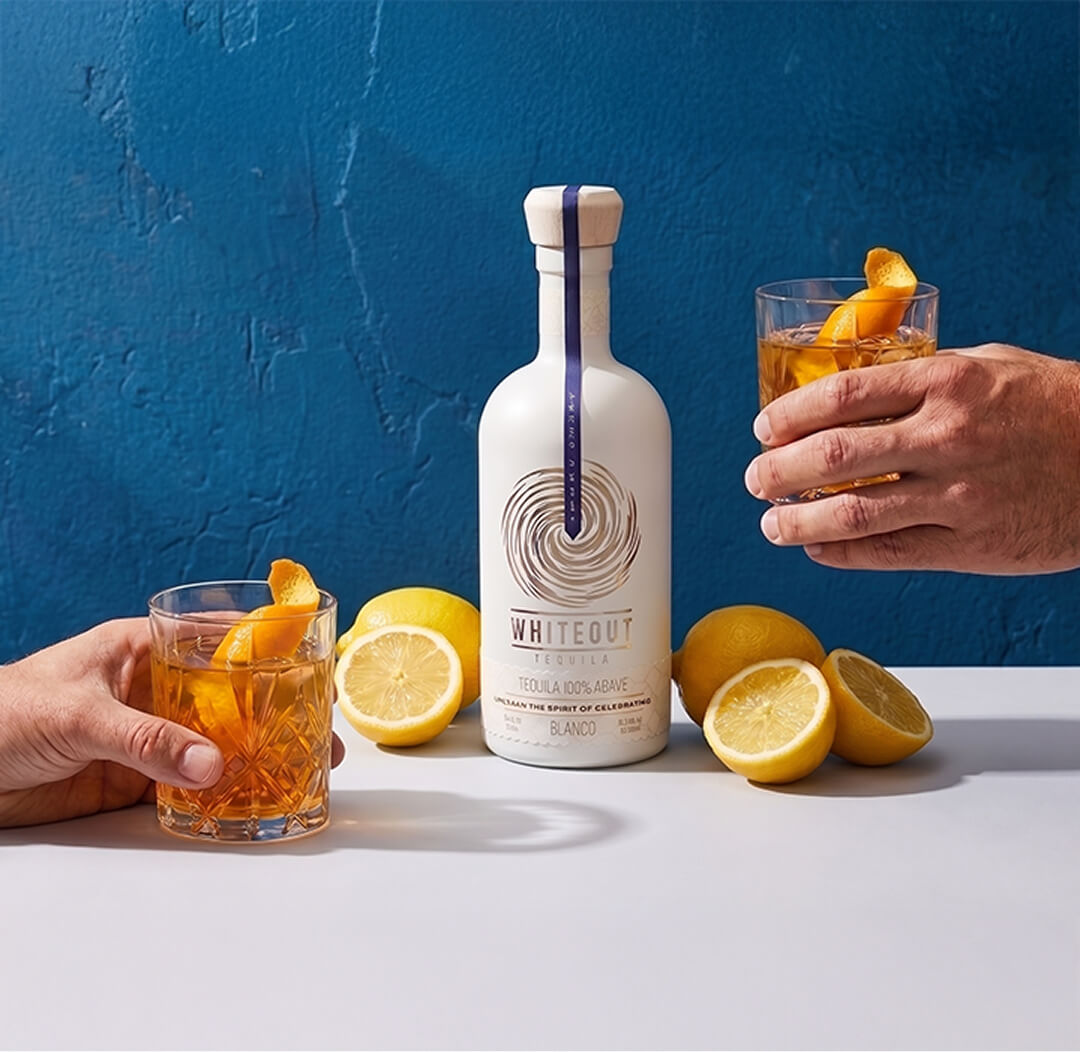 Whiteout Tequila Blanco served with two golden tequila cocktails and fresh lemon, styled against a bold blue backdrop.