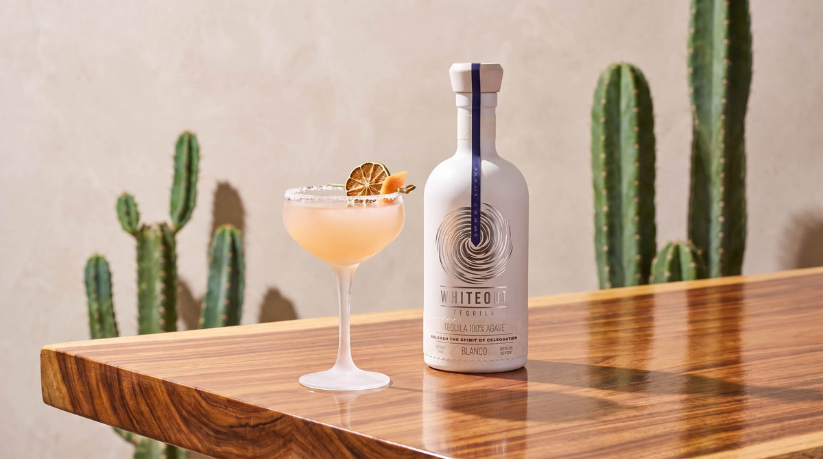 Whiteout Tequila Blanco bottle beside Jardín de Lychee cocktail on a wooden table with cactus plants in the backgroun